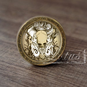 Könnte beinhalten: Ein runder, messingfarbener Knopf mit einem Totenkopf-Design. Der Totenkopf trägt eine Krone und ist von floralen Designs umgeben. Der Knopf ist perfekt, um jedem Raum einen Hauch von gotischem Stil zu verleihen.