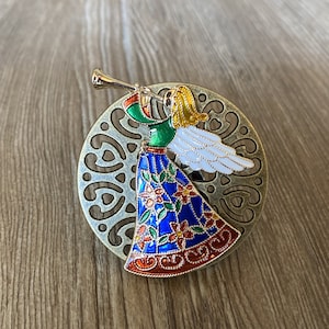 May include: A gold-coloured metal ornament featuring a colourful angel with blue, green, red, and white enamel paint. The angel is playing a trumpet and has a white halo. The ornament is set against a decorative, bronze-coloured background.