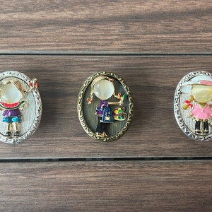 May include: Three oval-shaped brooches featuring cartoon girls with different outfits and accessories. The brooches have a silver, gold, and white metal finish.