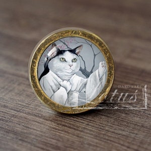 Peut inclure: Un bouton de porte rond en or avec un chat blanc portant une robe blanche et un visage noir et blanc. Le chat est devant un arbre avec des fleurs roses.