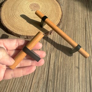 May include: Two wooden cabinet pulls with a light brown finish and black metal accents. One pull is held in a hand, while the other is on a wooden surface. The pulls have a simple, modern design.