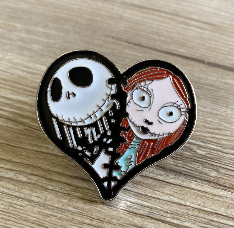 May include: A black and white heart-shaped pin with a cartoon image of Jack Skellington and Sally from the Nightmare Before Christmas movie. The pin is broken in half, with Jack on the left side and Sally on the right side.