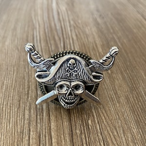 以下が含まれることがあります： 茶色の木製の背景に、交差した剣と三角帽子をかぶった銀の海賊の頭蓋骨