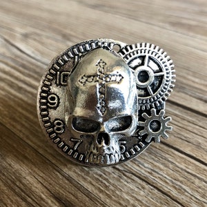 May include: Silver skull pendant with a cross design and gears. The skull has a clock face with Roman numerals.