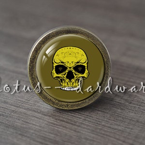 Puede incluir: Perilla de armario redonda de estilo antiguo con un diseño de calavera amarilla. La calavera tiene cuencas de ojos negras y un diseño amarillo detallado en la frente. La perilla tiene el texto "lotus-hardware" en cursiva.