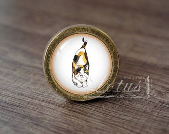 May include: A round, gold-toned cabinet knob with a white background and a watercolour illustration of a calico cat looking over its shoulder.
