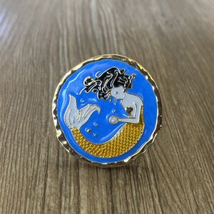 Könnte beinhalten: Eine goldfarbene Anstecknadel mit blauem Hintergrund, die eine Meerjungfrau mit schwarzen Haaren und einem gelben Schwanz zeigt. Die Meerjungfrau hält eine Perle.