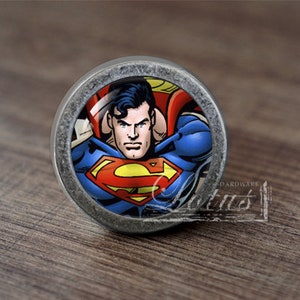 May include: A round, silver-colored cabinet knob with a Superman graphic. The graphic features Superman in his iconic red and blue suit with the Superman logo on his chest.