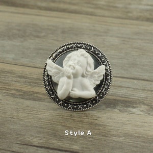 May include: A silver ring with a white cherub cameo. The cherub is depicted with wings and is holding its face. The ring has a decorative border with a cross pattern.