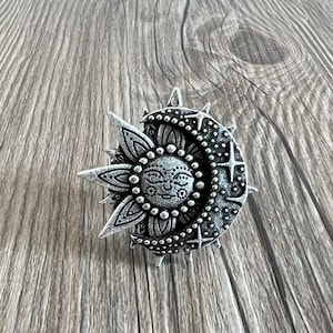 May include: A silver-toned pendant featuring a detailed sun and moon design. The sun has a face and is surrounded by rays, while the moon is crescent-shaped with stars.