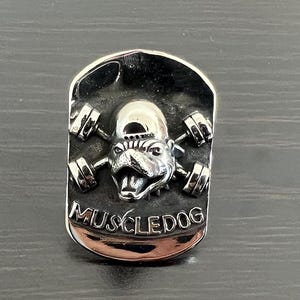 Könnte beinhalten: Silberfarbener, rechteckiger Anhänger mit einem Bulldoggen-Motiv, die einen Hut trägt und Langhanteln hebt. Der Text "MUSCLEDOG" ist unten eingraviert. Der Anhänger hat eine polierte, reflektierende Oberfläche.