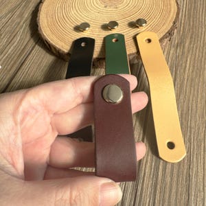 May include: A hand holds a burgundy leather key fob with a gold snap. Other key fobs in black, green, and yellow are visible, along with gold rivets and a wooden slice background. The key fobs are rectangular with rounded tops.