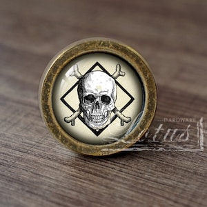 Könnte beinhalten: Ein runder, messingfarbener Schrankknopf mit einem Glaseinsatz, der eine Schwarzweißillustration eines Totenkopfs und gekreuzter Knochen in einem Diamanten zeigt.