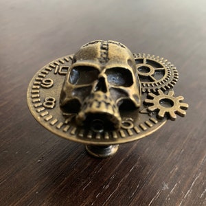 May include: A bronze skull with gears and a clock face, a decorative knob or pull for a cabinet or drawer.