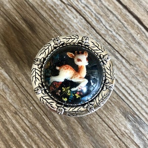 May include: A silver-toned round knob with a black background and a white deer illustration. The knob has a decorative border of leaves and flowers.