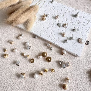 May include: A collection of decorative buttons in gold, silver, and pearl. The buttons are displayed on a textured white surface and a beige surface. Some buttons are attached to a white rectangular block. Several buttons have a pearl centre.