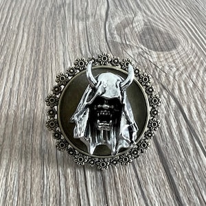 Puede incluir: Un colgante plateado con un demonio encapuchado con cuernos, sobre un fondo verde oscuro. El colgante está rodeado por un marco decorativo de color bronce con un diseño floral.