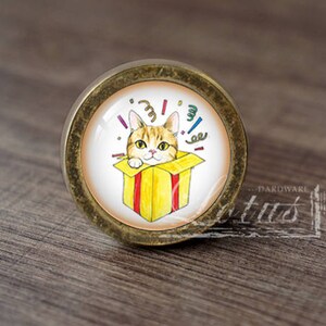 Może przedstawiać: Okrągły, złoty gałka meblowa z białym i żółtym wzorem. Kot wychyla się z żółtego pudełka z czerwonymi paskami. Konfetti otacza kota i pudełko.