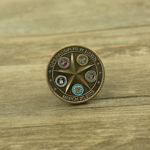 May include: A bronze-colored coin with a five-pointed star in the center. The star is surrounded by the seals of the five branches of the United States Armed Forces. The coin is inscribed with "Operation New Dawn" and "March 20, 2003".