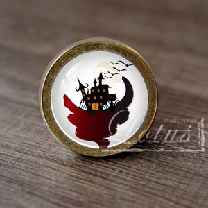 Pode incluir: Um botão de armário redondo de cor latão com um inserto de cerâmica branca que apresenta uma silhueta preta de uma casa assombrada, uma lua crescente e morcegos sobre um fundo branco.