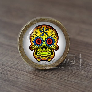 Puede incluir: Un pomo de armario redondo de color latón con un fondo blanco y un diseño de calavera de azúcar colorido. La calavera tiene detalles amarillos, naranjas, rojos, azules y verdes.