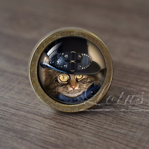 Op de afbeelding: Een ronde, messingkleurige kastknop met een glazen inzetstuk met een kat die een zwarte hoge hoed draagt met steampunk-accenten en een monocle.