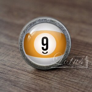 Op de afbeelding: Een zilveren knop met een ronde glazen inzet met een gele en witte biljartbal met het nummer 9.
