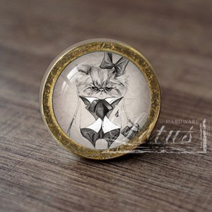 Op de afbeelding: Een ronde, gouden kastknop met een glazen inzetstuk met een zwart-witte illustratie van een kat in smoking met een strikje.