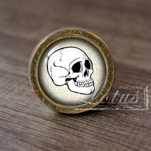 Puede incluir: Un pomo de armario redondo de color dorado antiguo con un diseño de calavera blanca sobre un fondo crema. La calavera tiene ojos negros y una mandíbula detallada.