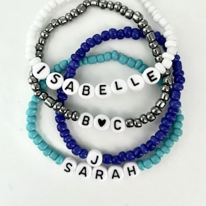 May include: Four beaded bracelets with the names "Isabelle", "B♥C", and "Sarah" spelled out in white beads with black lettering. The bracelets are layered in blue, turquoise, silver, and white.