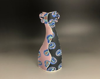 May include: A ceramic vase with a unique, abstract design. The vase is black and pink with blue and brown accents. The vase has a narrow neck and a wide base.