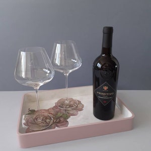 May include: A pink serving tray with a floral design featuring roses. Two clear wine glasses and a bottle of wine labelled "Primitivo" are on the tray.
