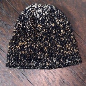 May include: A black, white, and brown knit beanie hat. The hat is made of a soft, textured yarn.
