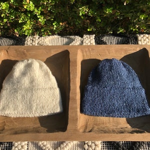 May include: Two knitted hats, one white and one blue, are displayed in a wooden tray. The hats are made of a soft, textured yarn.