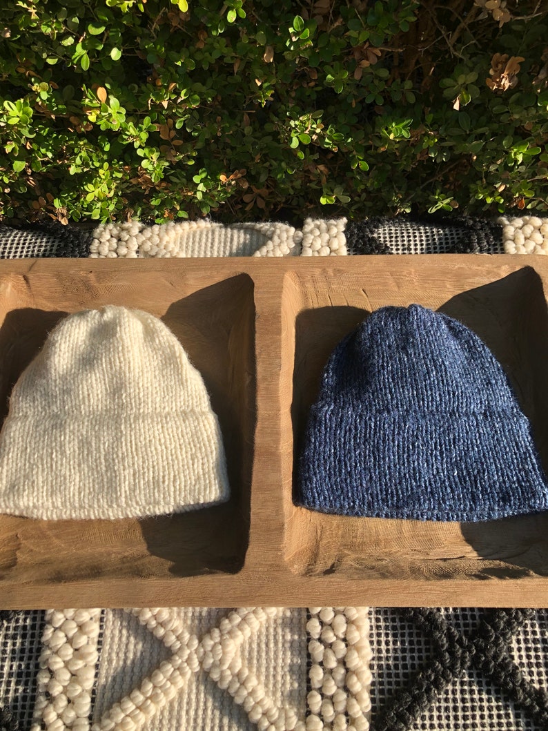 May include: Two knitted hats, one white and one blue, are displayed in a wooden tray. The hats are made of a soft, textured yarn.