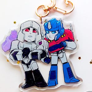 May include: A 2.5 inch acrylic charm featuring Optimus Prime and Megatron from the Transformers franchise. The charm is clear with a glittery background and features the characters in a chibi style. The charm is attached to a gold-colored key chain.
