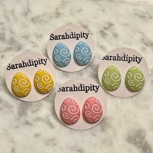 May include: Four pairs of Easter egg-shaped earrings. Each pair is a different color: blue, yellow, green, and pink. The earrings have a white swirl design.