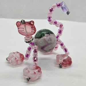 May include: A beaded cat figurine with a pink and purple color scheme. The cat's head is a pink cat-shaped bead, and its body is a large, round, speckled stone. The legs and tail are made of pink and purple beads.