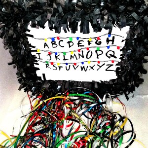 May include: A black and white pinata shaped like the head of a character from the show Stranger Things. The pinata has the alphabet written on it with colorful light bulbs. The pinata is surrounded by colorful streamers.