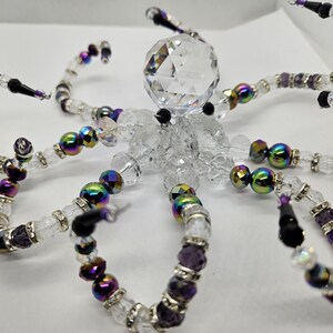 May include: A decorative crystal octopus sculpture. The sculpture features a large, faceted crystal head and body, with eight articulated tentacles. The tentacles are made of clear and purple beads, with iridescent and black accents. The sculpture is on a white background.