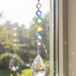 May include: A decorative suncatcher with a chain and a teardrop-shaped crystal pendant. The chain holds a series of colorful, round glass beads in blue, green, yellow, and pink. The suncatcher hangs in front of a window with a blurred outdoor background.
