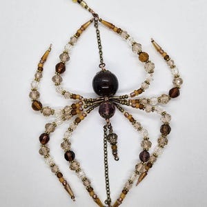 May include: A handcrafted spider-shaped ornament made of brown, amber, and clear beads. The spider has eight beaded legs, a large central bead, and a decorative wirework head. A chain and additional beads hang from the spider's body.