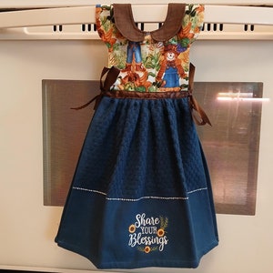 May include: A blue kitchen towel with a brown ribbon and a brown and white fabric top with a scarecrow design. The towel has the text "Share Your Blessings" embroidered on it with sunflowers.
