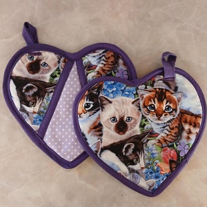 May include: Two purple heart-shaped pot holders with a white polka dot pattern. The pot holders are decorated with a colorful print of cats.