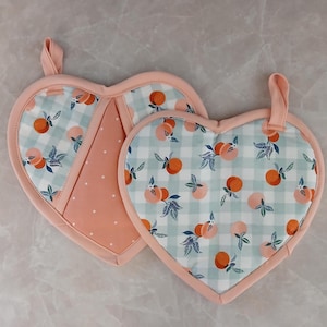 May include: Two heart-shaped pot holders. One features a light blue and white gingham pattern with orange peaches and green leaves. The other has a peach-coloured polka dot pattern. Both have peach-coloured trim and hanging loops.