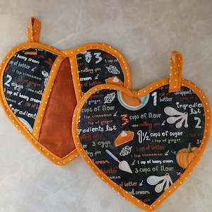 May include: Two heart-shaped orange pot holders with black and white text and illustrations. The text includes a recipe for pumpkin pie with ingredients and measurements. The pot holders are ideal for baking and cooking.