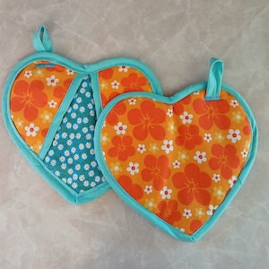 以下が含まれることがあります： ハート型の鍋つかみ2つ。オレンジ色の花柄と白い花の模様、もう一つはターコイズとオレンジのデザイン。どちらもターコイズの縁取りと吊り下げループが付いています。キッチン装飾。