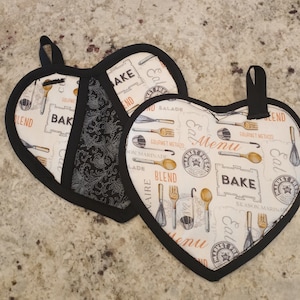 Puede incluir: Dos agarraderas de olla en forma de corazón con ribete negro, fondo blanco y diseño negro y marrón con utensilios de cocina, texto que dice "Market Fresh", "Menu", "Bake", "Blend" y "Gourmet Method".