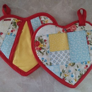 Puede incluir: Dos agarraderas de olla en forma de corazón con ribete rojo y diseño de patchwork. Las agarraderas están hechas de una variedad de telas en tonos de amarillo, azul y blanco con patrones florales.
