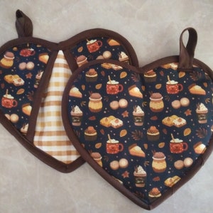 Könnte beinhalten: Zwei herzförmige Topflappen mit braunem Rand. Ein Topflappen ist in Braun und Weiß kariert, der andere ist dunkelblau mit einem Muster aus braunen und orangen Tassen, Cupcakes und Tortenstücken.